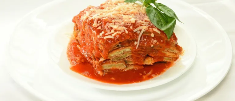 A square piece of lasagna sprinkled with cheese on a plate with a basil leaf garnish.