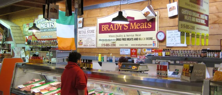 A Brady's staff member conversing with a customer in front of the Brady's Meat & Deli stall at the St. Jacobs Farmers Market