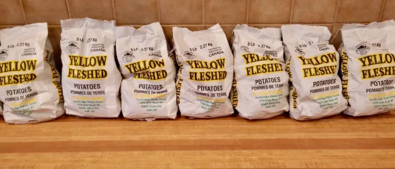 Seven bags of Canadian, yellow-fleshed potatoes in a line on a wooden counter.
