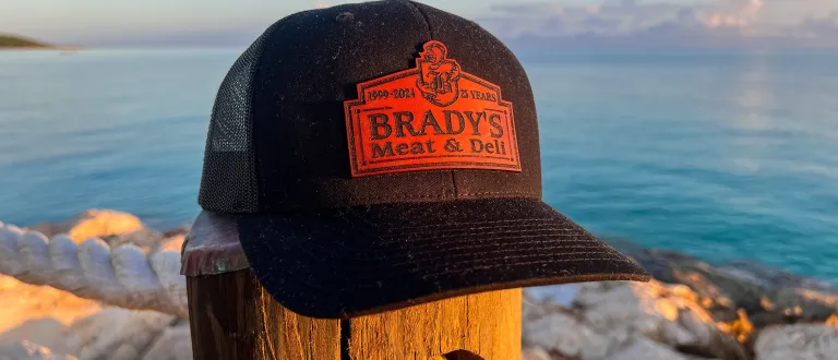 A black, mesh baseball cap with the Brady's Meat & Deli logo resting on a fencepost on a stony beach.